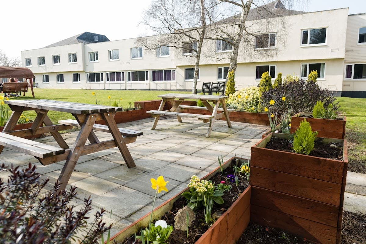 Outdoor seating area with wooden picnic tables, flower beds, and a modern hotel building in the background.