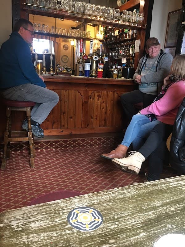 Cozy bar interior with patrons chatting, wooden bar, various drinks displayed, and patterned carpet.