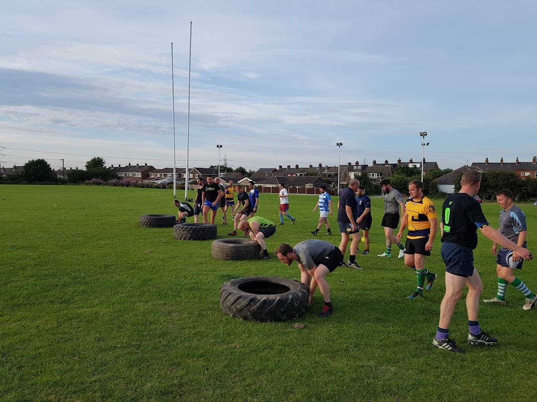 Rugby players training on a grassy field, using large tires for drills, with goalposts in the background.