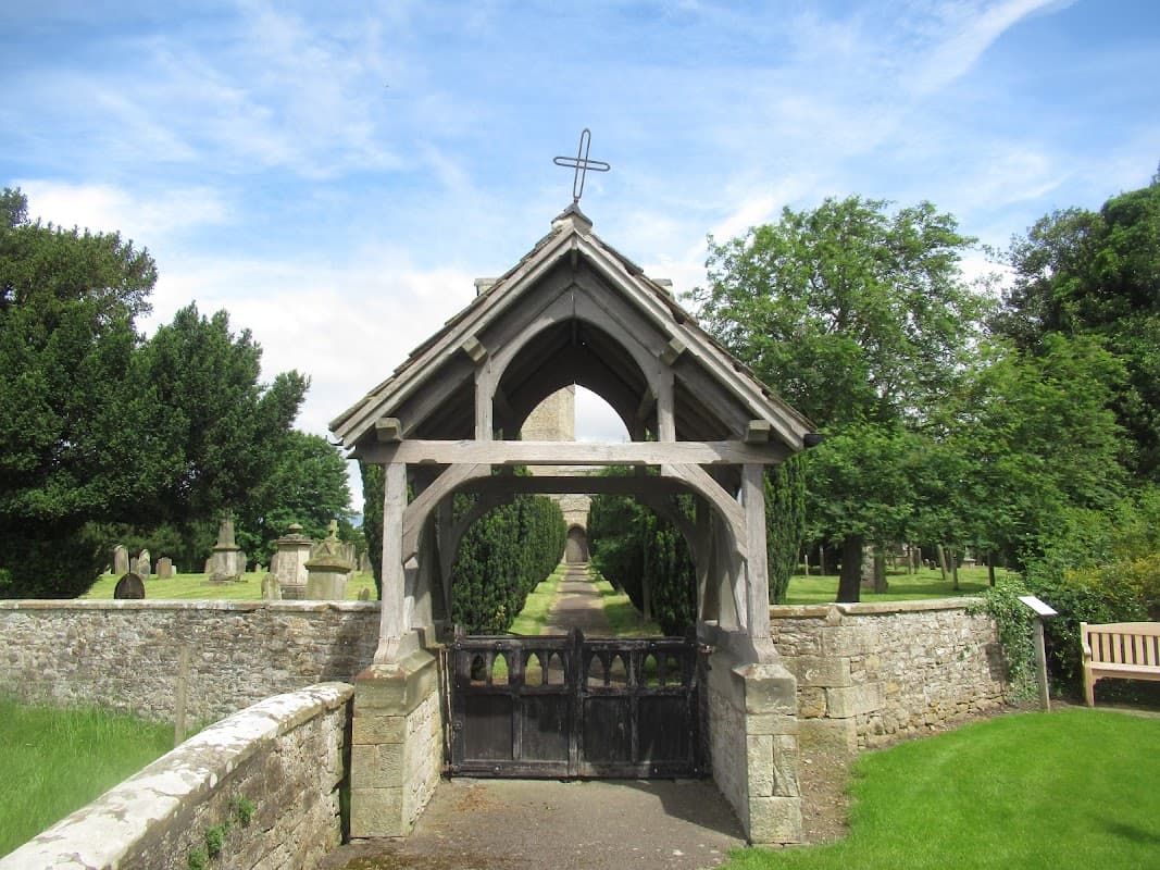 Wooden entrance with a cross, leading to a church surrounded by trees and gravestones in a serene landscape.