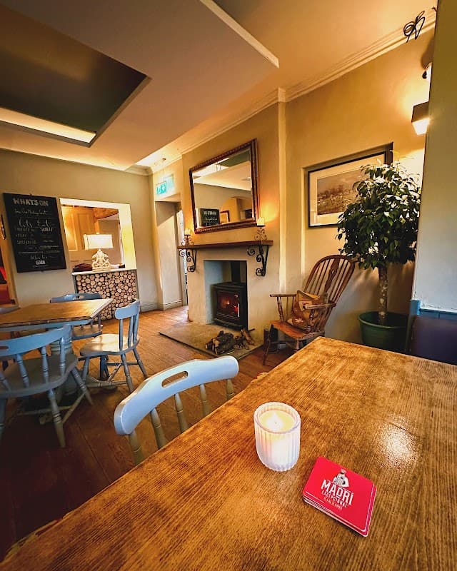 Cozy interior of The Stanwick Arms with a wooden table, candle, armchair, and fireplace, creating a warm atmosphere.