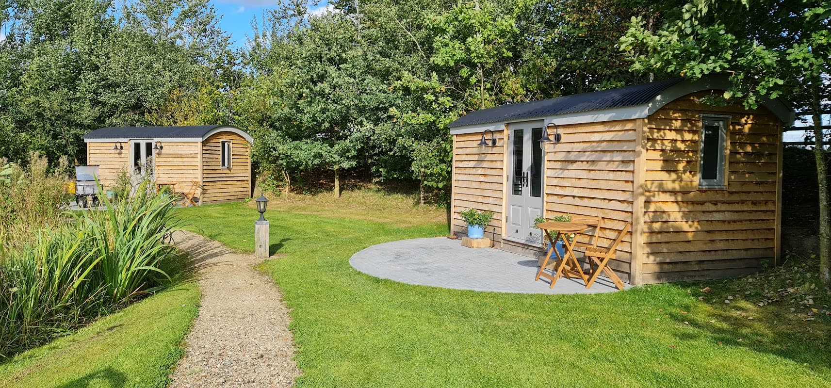 Two wooden cabins in a grassy area surrounded by trees, with a stone path and outdoor seating.