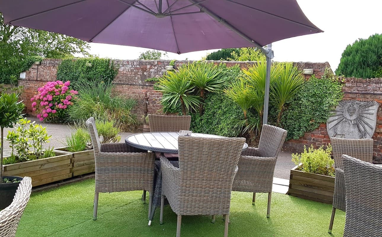 Outdoor seating area with wicker chairs, a round table, and vibrant flowers against a rustic brick wall.