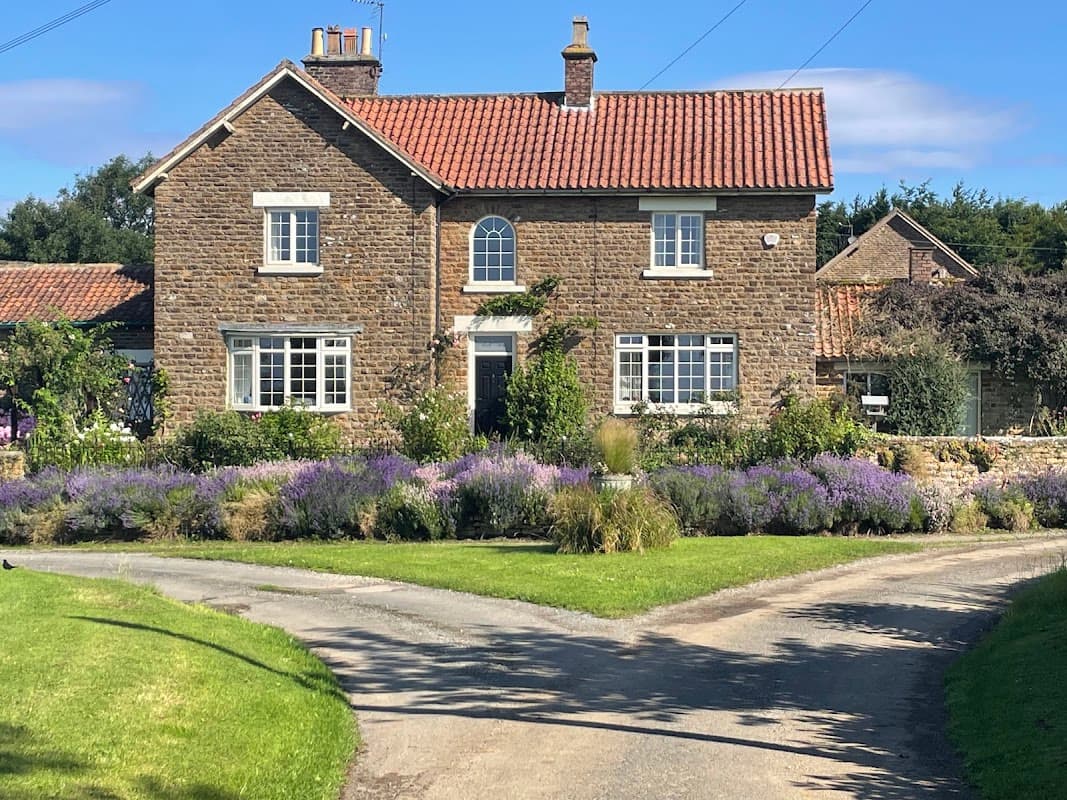 Charming stone house with red-tiled roof, surrounded by lush greenery and lavender flowers, set on a winding driveway.