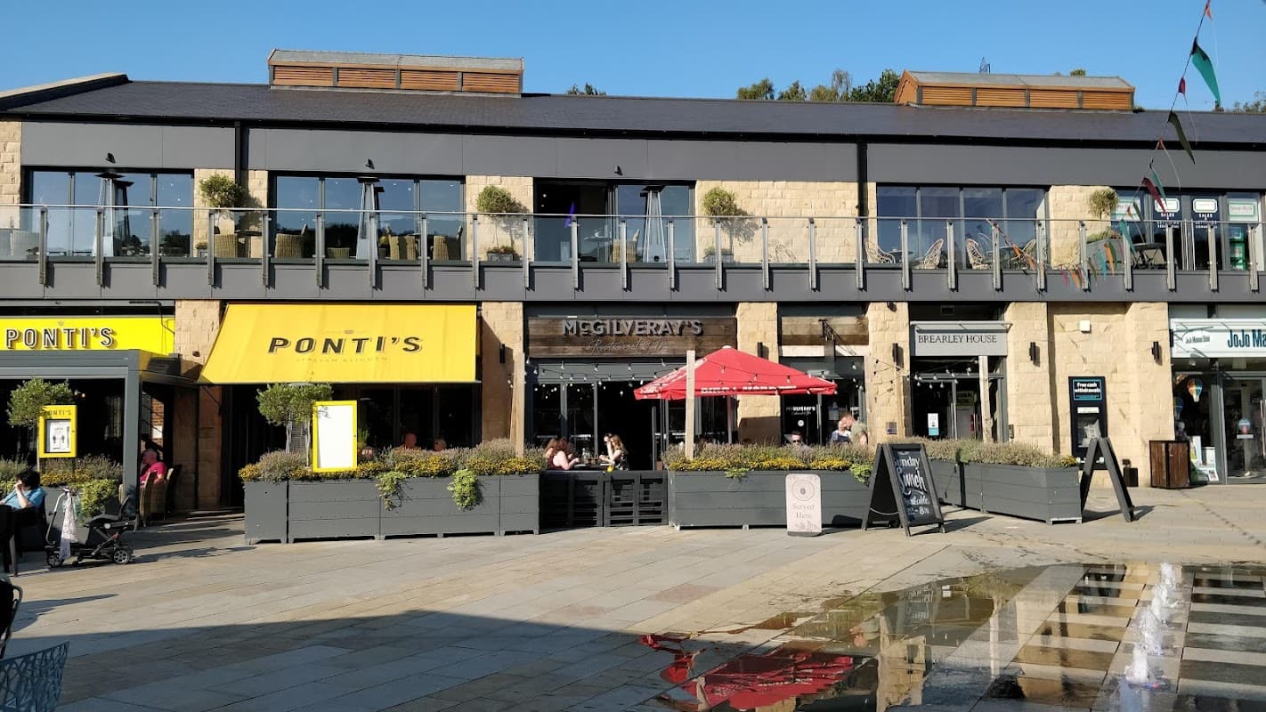 Shopping center featuring restaurants with outdoor seating, planters, and a sunny atmosphere in Stocksbridge, Yorkshire.