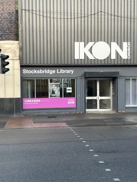 Stocksbridge Library storefront with large windows, a pink sign, and the adjacent IKON Church building.