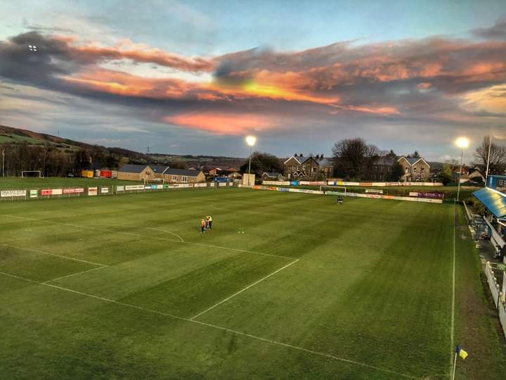 Football pitch at Stocksbridge Park Steels FC under a colorful sunset, with players warming up and floodlights on.