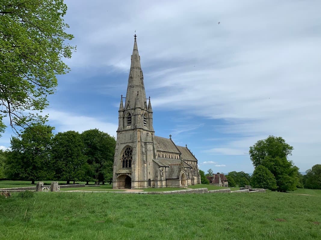 Gothic-style church with a tall spire surrounded by lush greenery and blue sky at Studley Royal Deer Park.