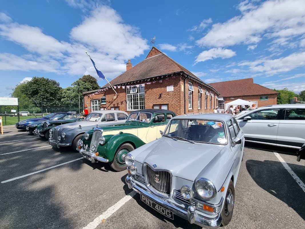 Sutton Upon Derwent Village Hall with classic cars parked in front under a blue sky with scattered clouds.
