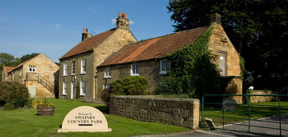 Stone buildings with red roofs, green grass, and a welcoming sign for Swainby Country Park in a sunny landscape.