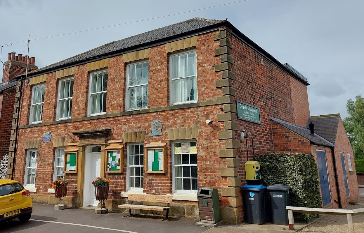 Red brick building with large windows, a bench outside, and signs for Swainby Village Hall. Green bins nearby.