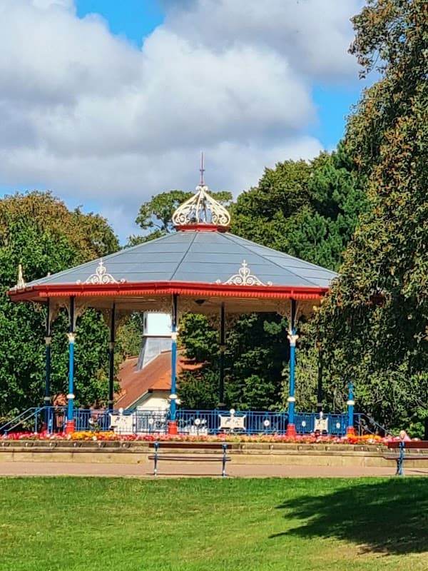 Victorian bandstand surrounded by trees and colorful flower beds under a blue sky with fluffy clouds.