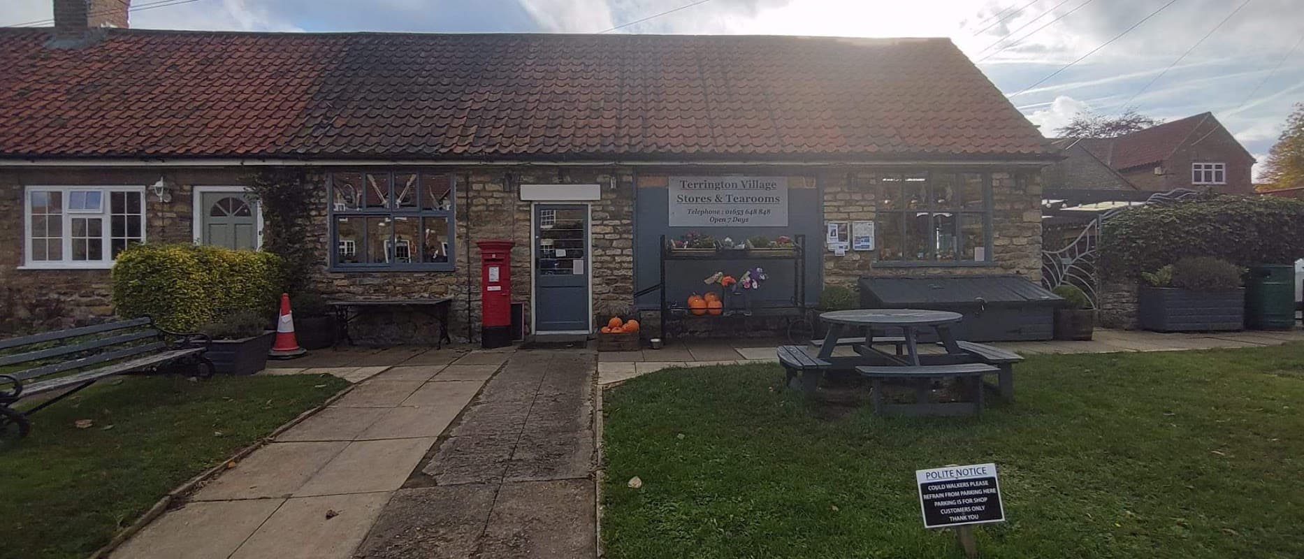 Quaint stone building with a red post box, outdoor seating, pumpkins, and a sign for Terrington Village Store.