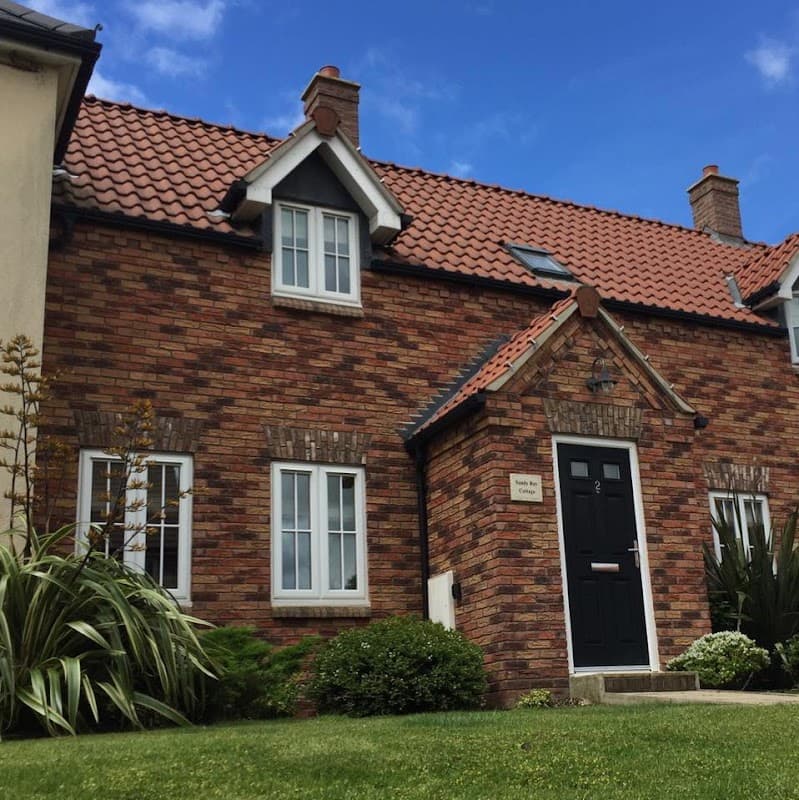 Charming brick cottage with a black door, white windows, and lush greenery under a blue sky in Sandy Bay, Filey.