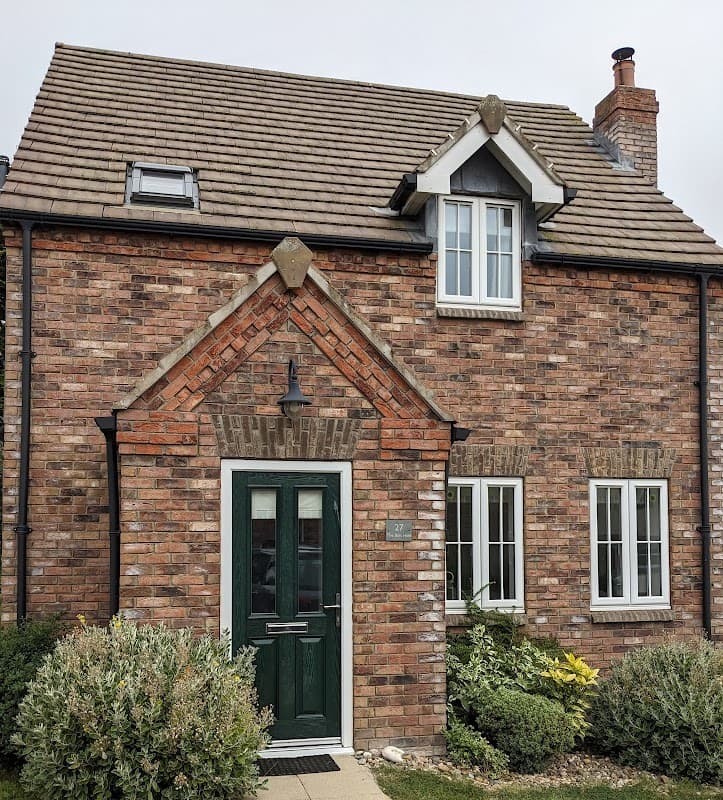 Charming brick cottage with a green door, white windows, and a slate roof, surrounded by lush greenery.