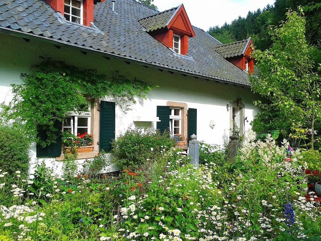Charming farmhouse with red accents, surrounded by a lush garden filled with flowers and greenery.