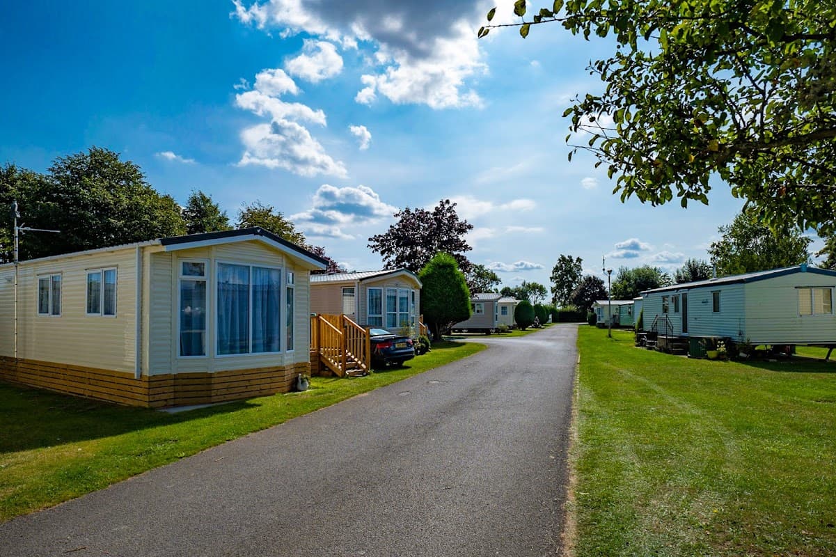 Caravans lined along a paved path in a green holiday park under a blue sky with scattered clouds.