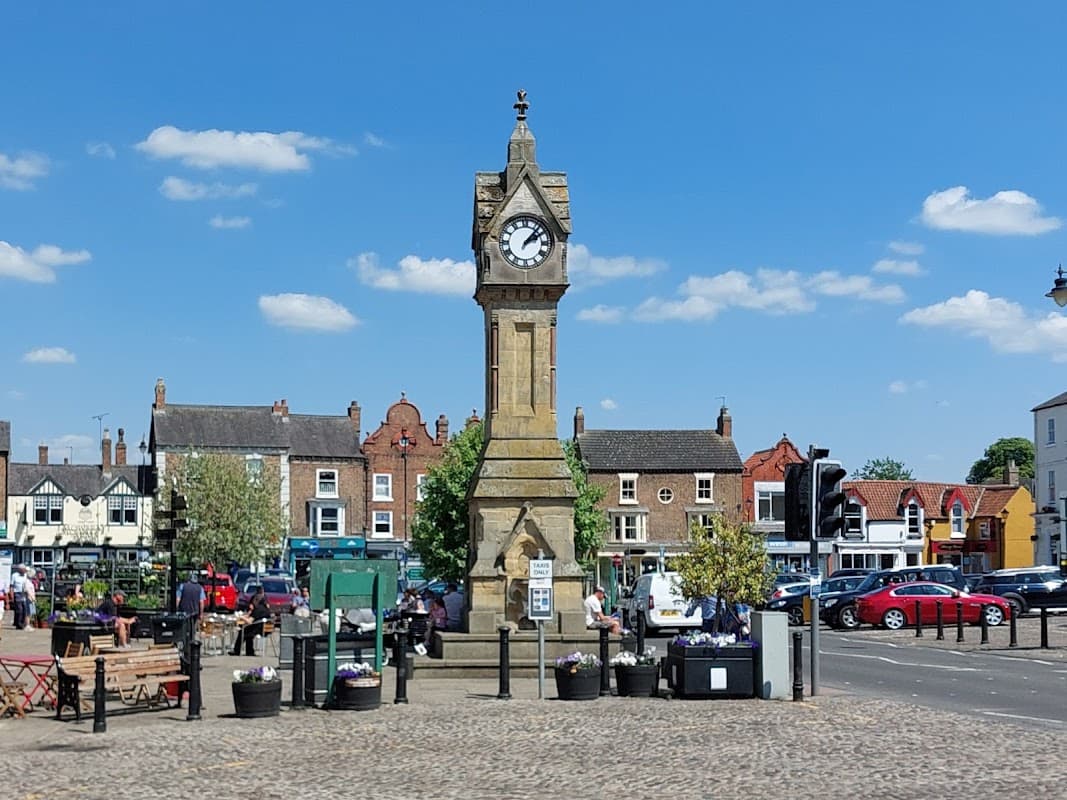 Historic clock tower in a bustling market square, surrounded by shops, greenery, and parked cars under a clear blue sky.