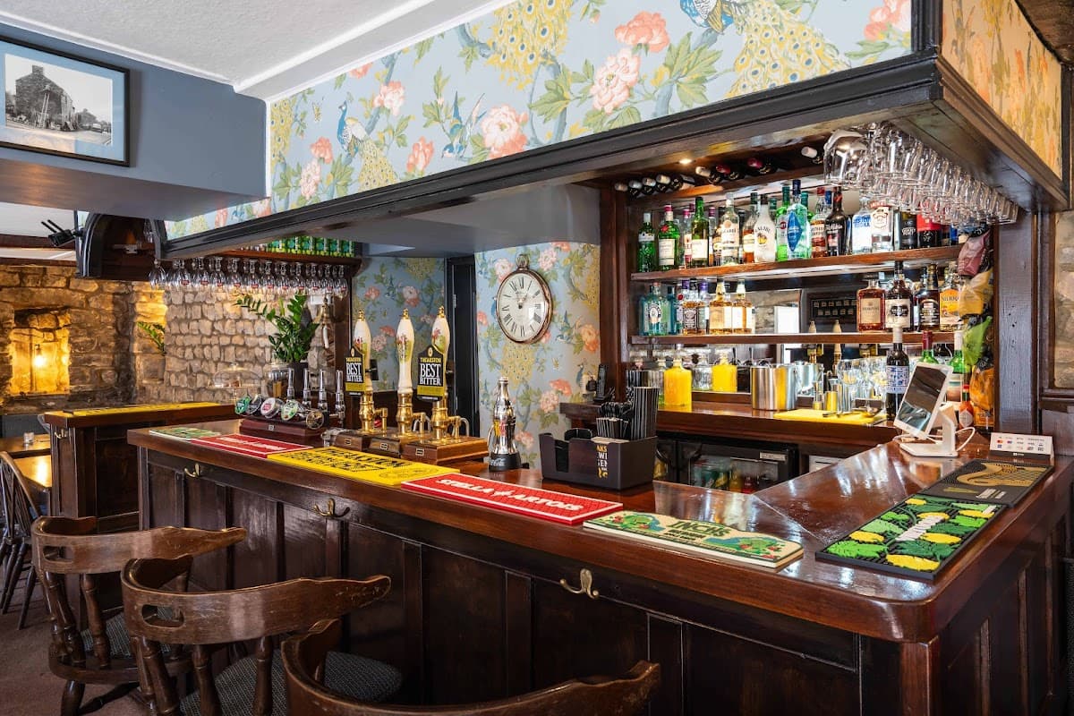 Wooden bar with floral wallpaper, shelves of drinks, clocks, and bar stools in a cozy pub setting.