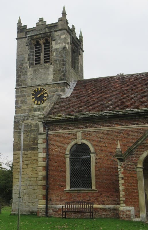 Church tower with clock, stone and brick walls, and a bench in front, set in a grassy area in Thorganby, North Yorkshire.
