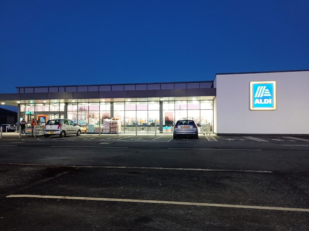 ALDI store exterior at dusk, featuring large windows and a prominent logo, with parked cars in the foreground.