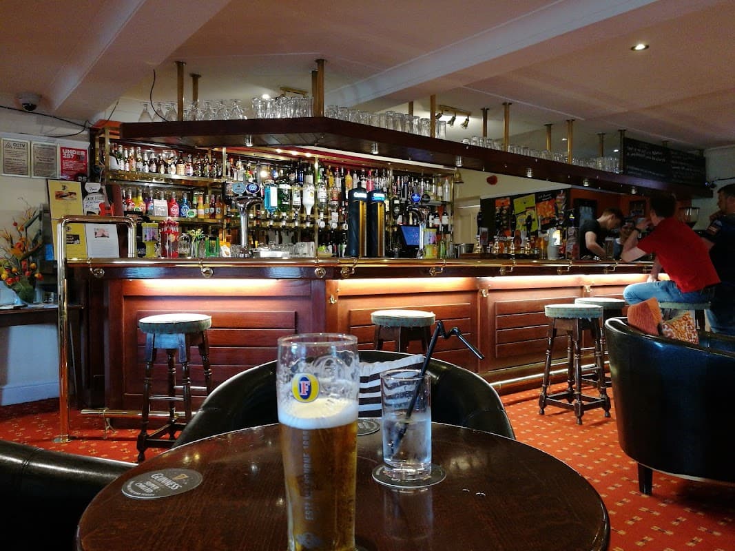 Wooden bar with various drinks, bar stools, and tables; patrons chatting in a cozy, warmly lit atmosphere.