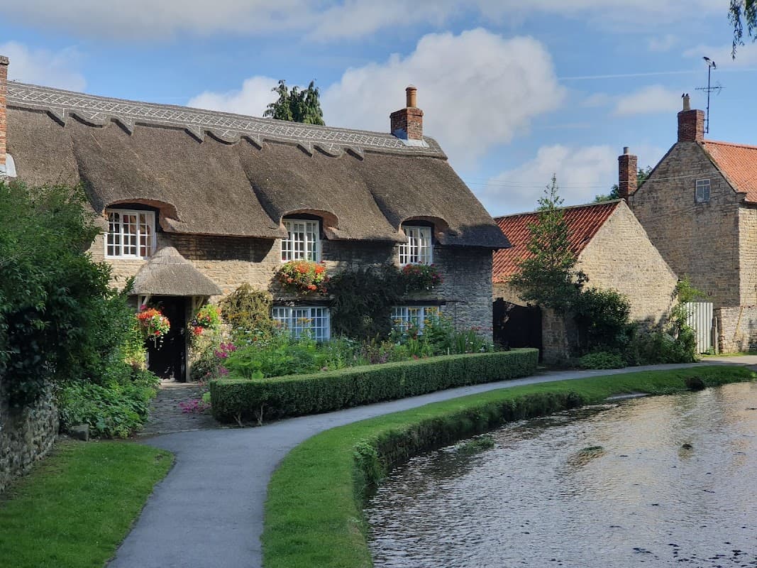 Thatched cottage with flower boxes, lush greenery, and a tranquil stream in Thornton-le-Dale, Yorkshire.
