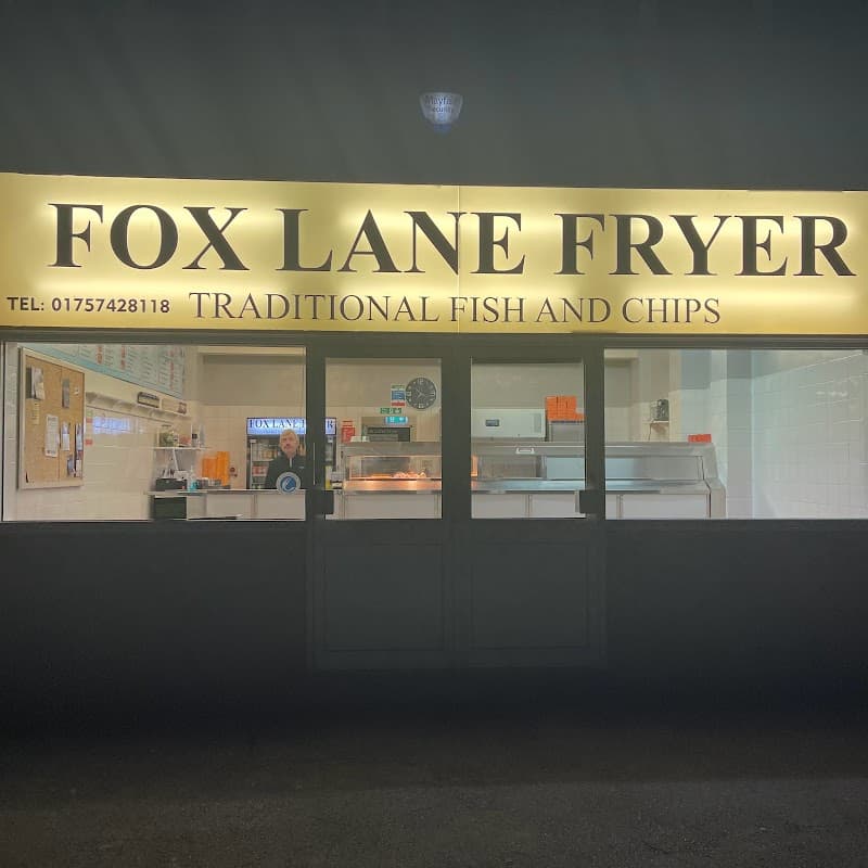 Brightly lit restaurant sign for Fox Lane Fryer, featuring traditional fish and chips, with a staff member visible inside.