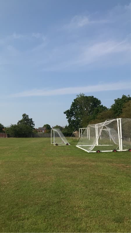 Football goals on a grassy field under a clear blue sky, surrounded by trees and distant buildings.