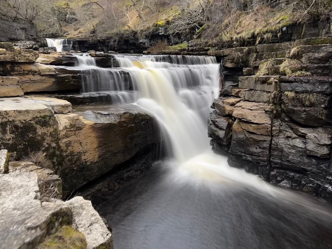 A cascading waterfall flows over rocky ledges in a serene landscape surrounded by trees and mossy stones.