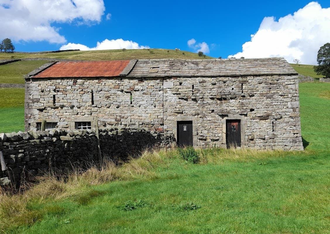 Stone building with a red roof, surrounded by green grass and dry stone walls, under a blue sky with fluffy clouds.