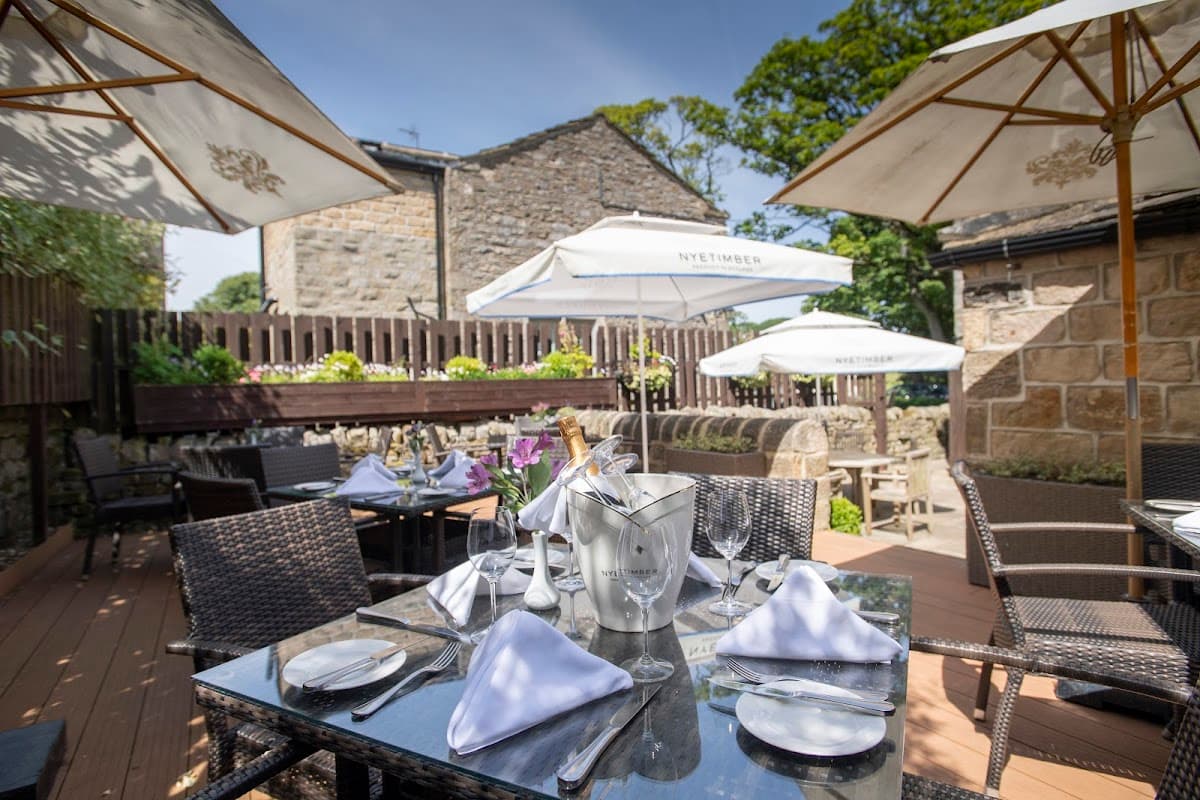 Outdoor dining area with a table set for guests, umbrellas, and stone buildings in a lush garden setting.