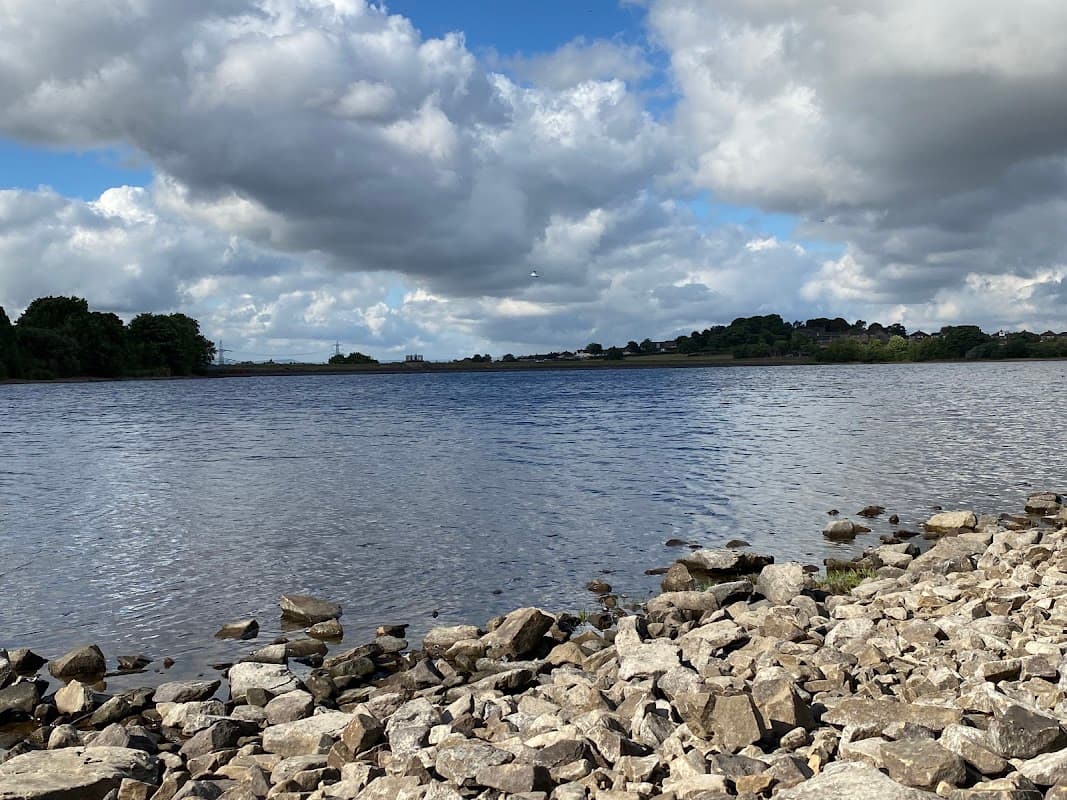 Calm water of Ardsley Reservoir surrounded by rocky shoreline and lush greenery under a cloudy blue sky.
