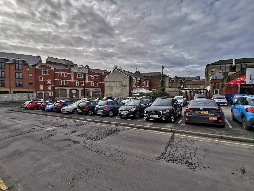Free parking lot in Tingley, Yorkshire, with various parked cars and buildings in the background under a cloudy sky.