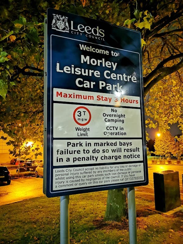 Sign for Morley Leisure Centre Car Park, detailing stay limits, weight restrictions, and CCTV operation. Trees in background.