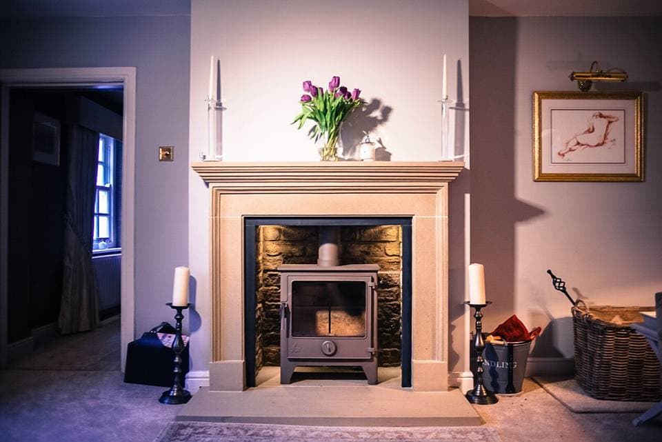 A cozy living room featuring a stone fireplace, a wood stove, candles, and a vase of tulips on the mantel.