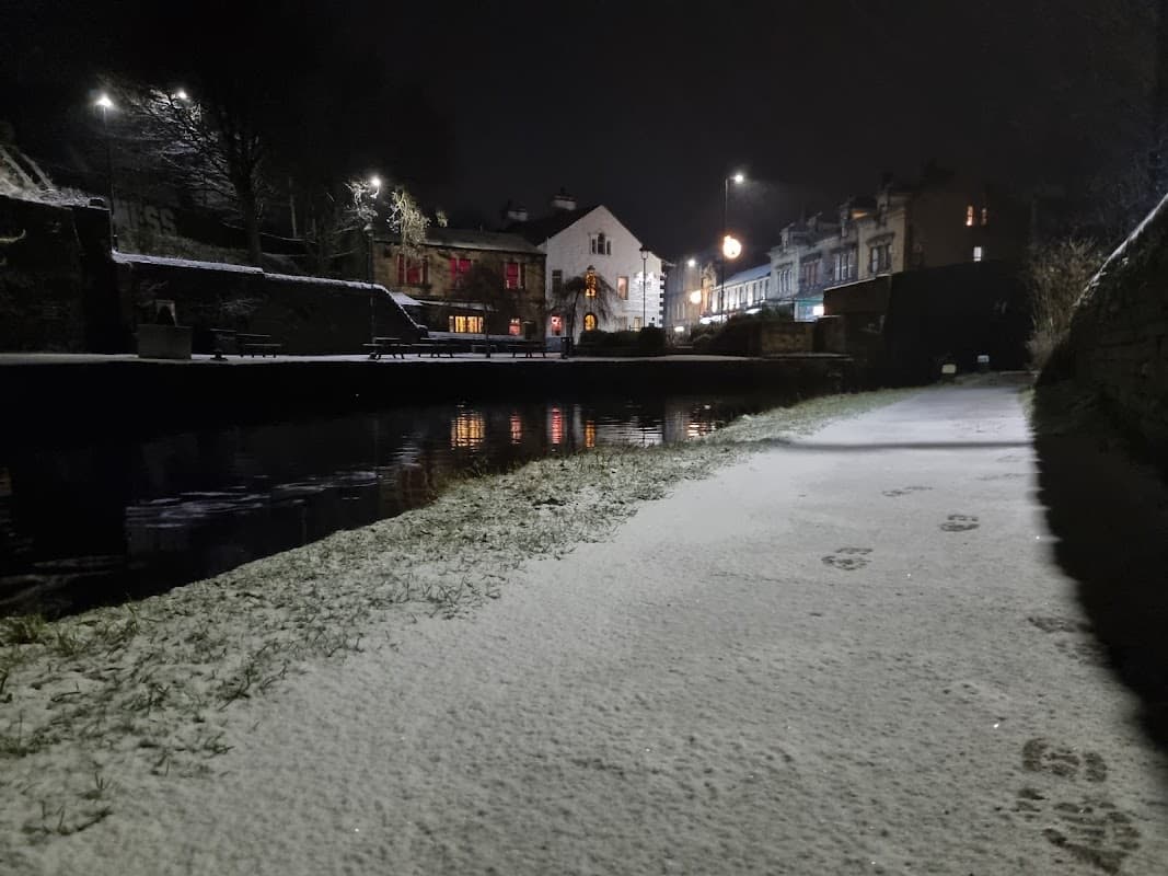 Snow-covered path beside a canal at night, with illuminated buildings and trees in the background.