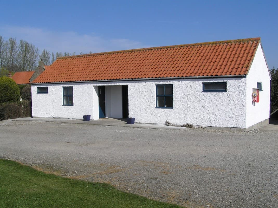 White building with a red-tiled roof, surrounded by a gravel area and green grass under a clear blue sky.