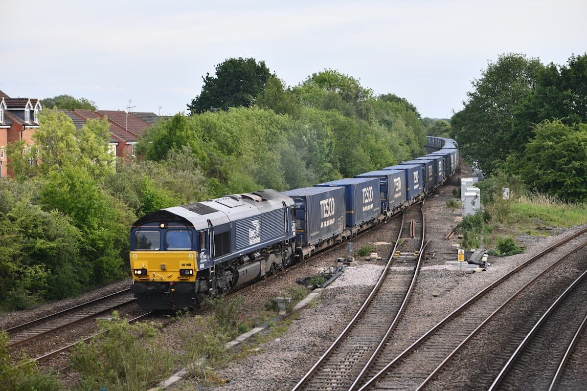 A freight train with blue containers passes through Church Fenton Station, surrounded by greenery and railway tracks.