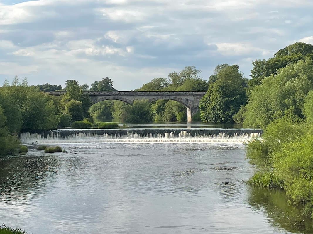 A serene river scene with a stone bridge and lush greenery lining the banks, under a cloudy sky.