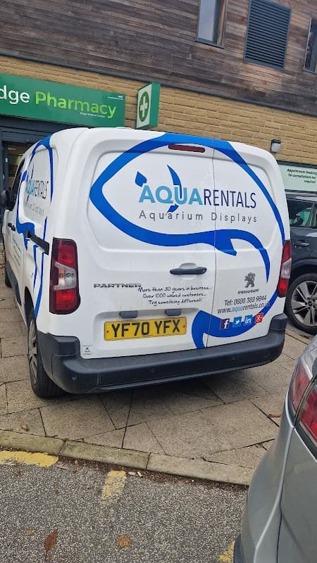 White van with "Aqua Rentals" logo, featuring blue waves and text about aquarium displays, parked near a pharmacy.