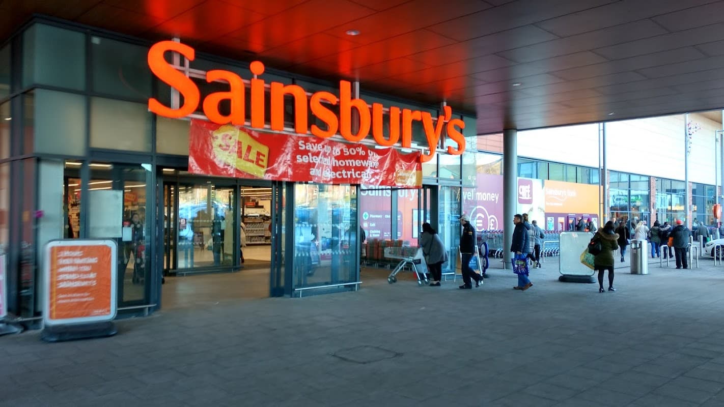 Sainsbury's storefront with large red signage, shoppers entering and exiting, and various promotional banners visible.