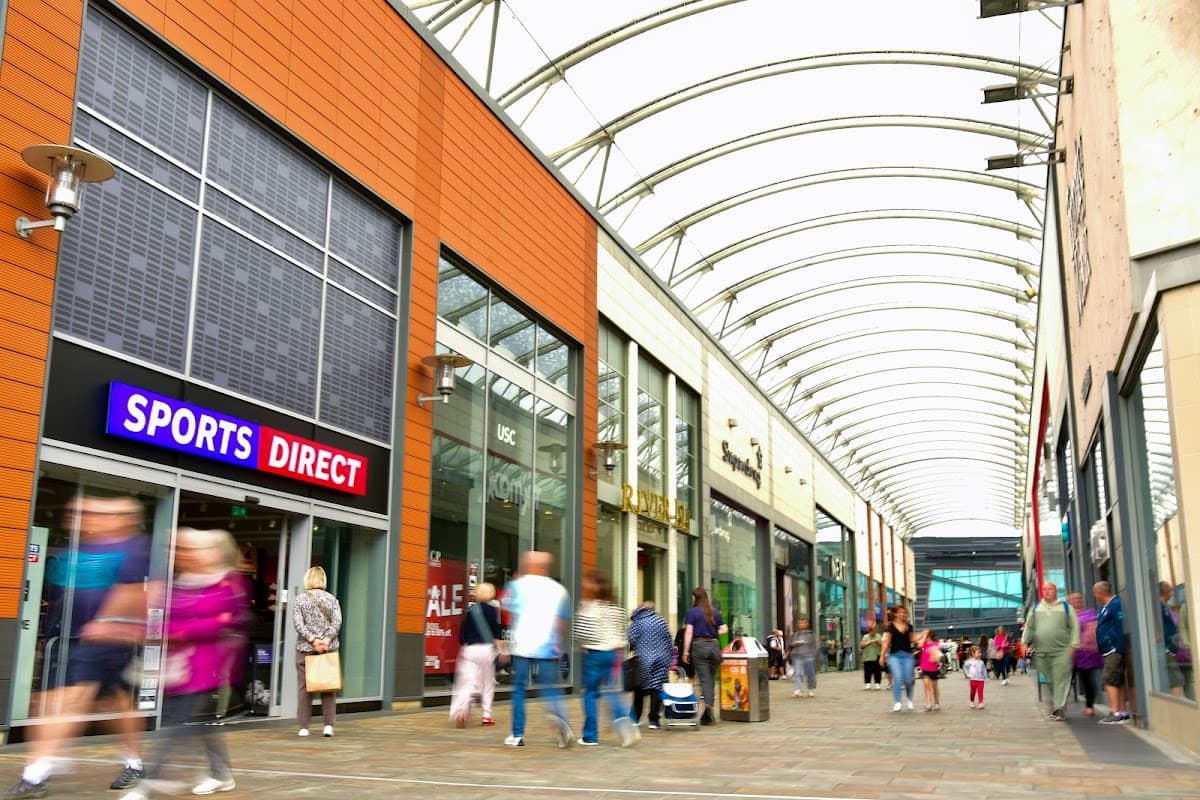 Busy shopping arcade with a Sports Direct store, modern architecture, and shoppers walking under a curved glass roof.