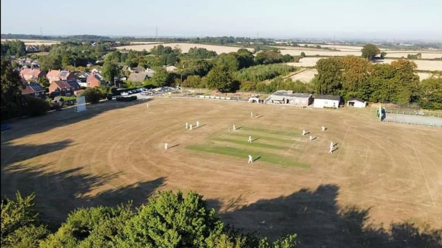Aerial view of Walkington Cricket Club with players on a dry pitch, surrounded by trees and houses in the background.