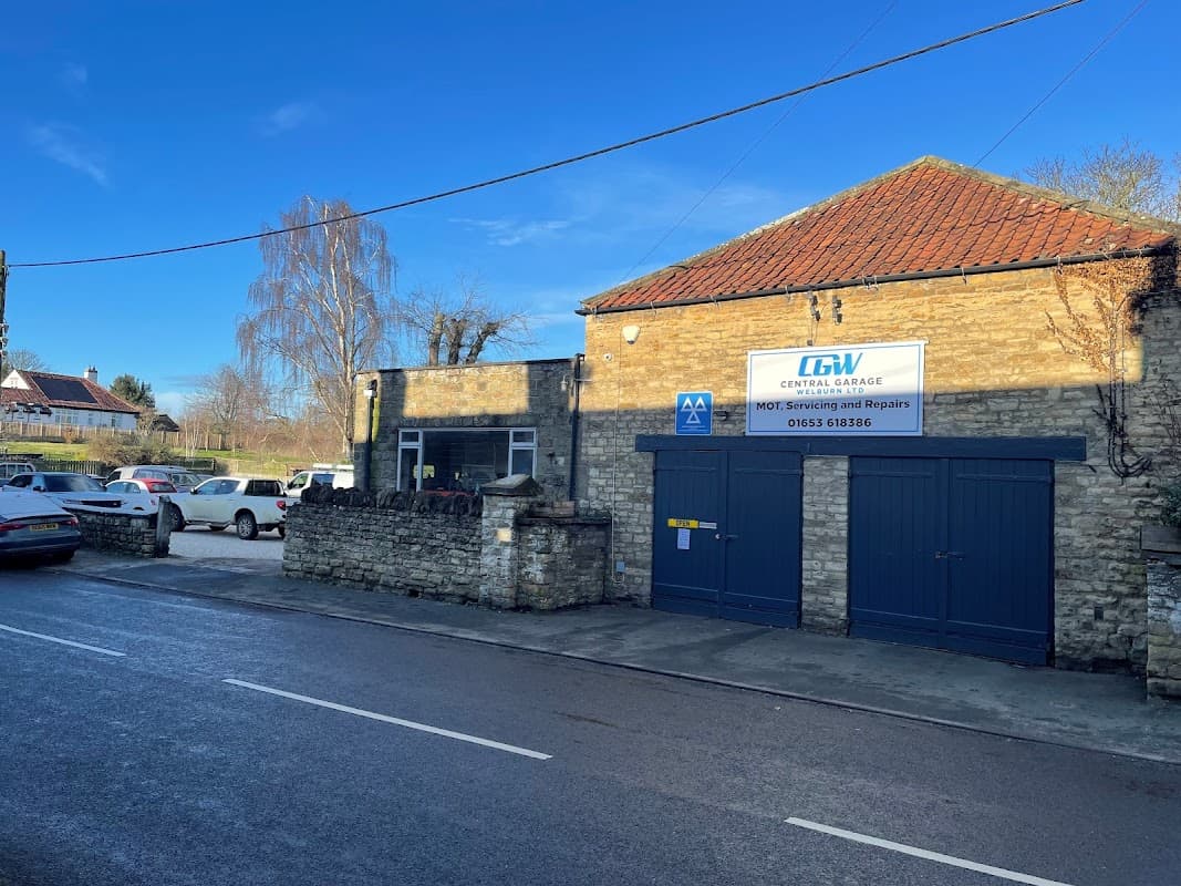 Central Garage Welburn Ltd building with stone walls, blue garage doors, and a sign on a clear, sunny day.