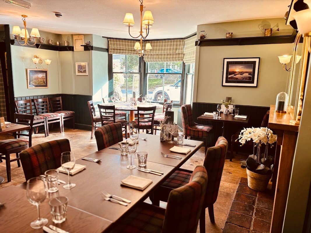 Cozy restaurant interior with wooden tables, plaid chairs, and soft lighting, featuring floral decor and scenic wall art.