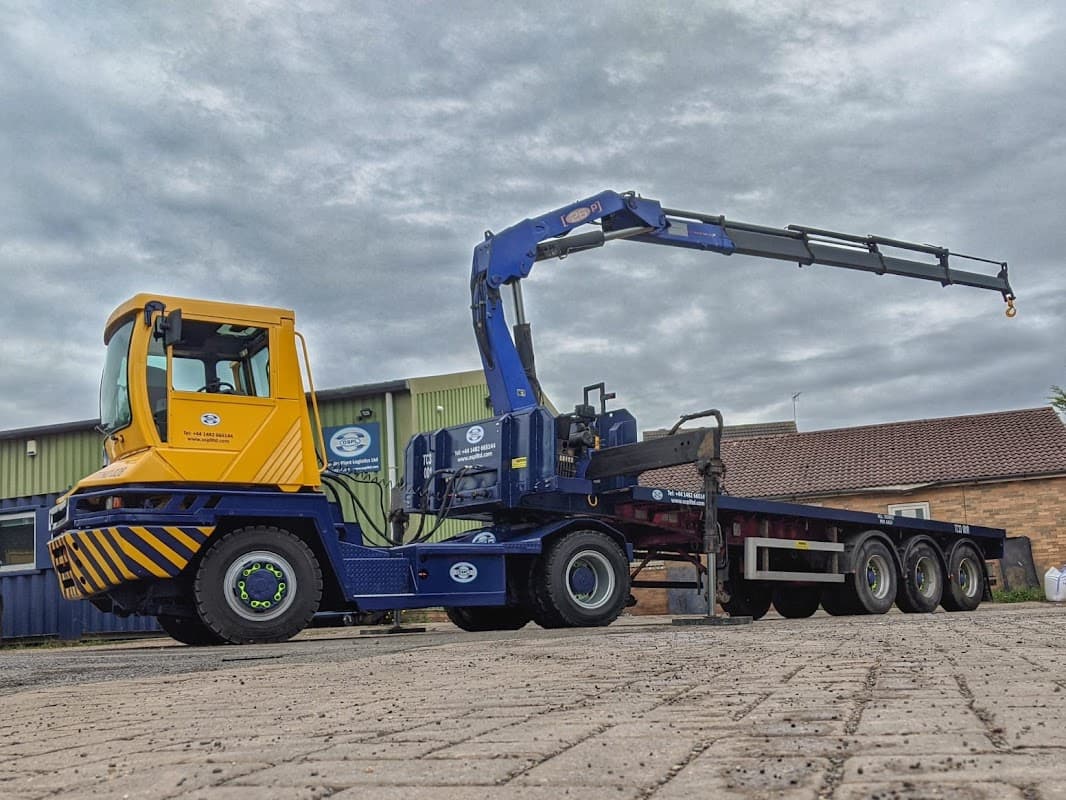 Yellow and blue truck with a crane attachment parked on a paved area, surrounded by industrial buildings and cloudy sky.