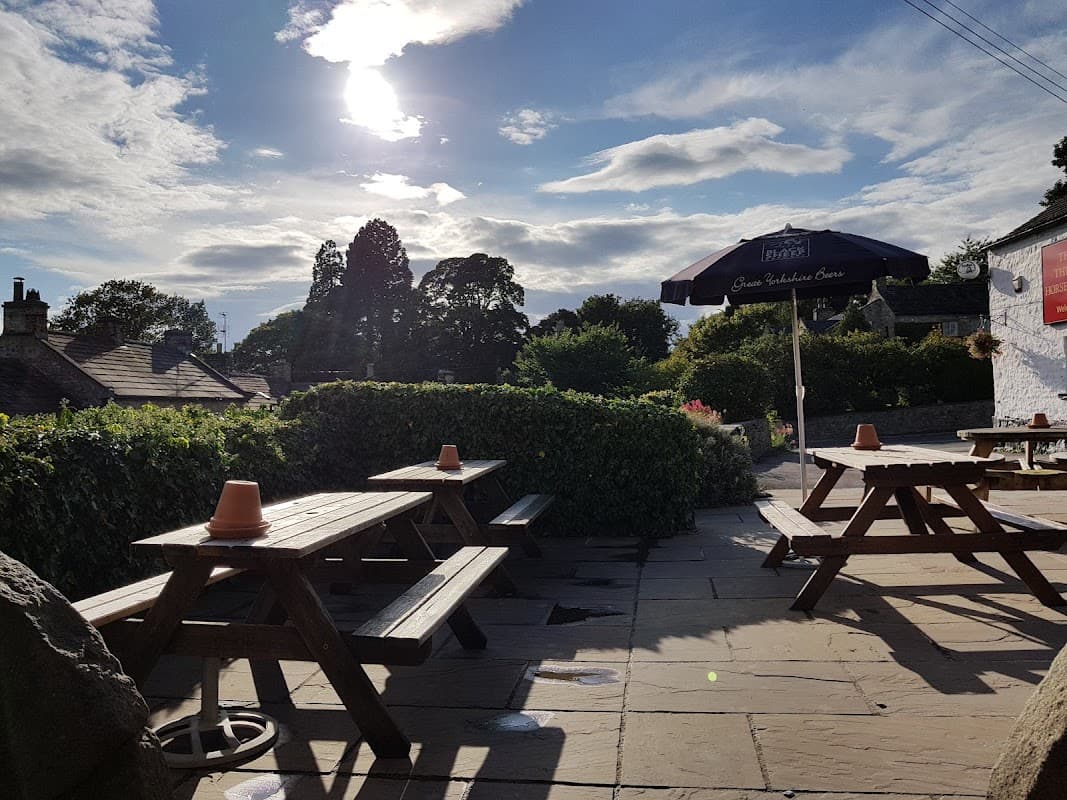 Outdoor seating area with wooden picnic tables, surrounded by greenery, under a sunny sky with clouds.