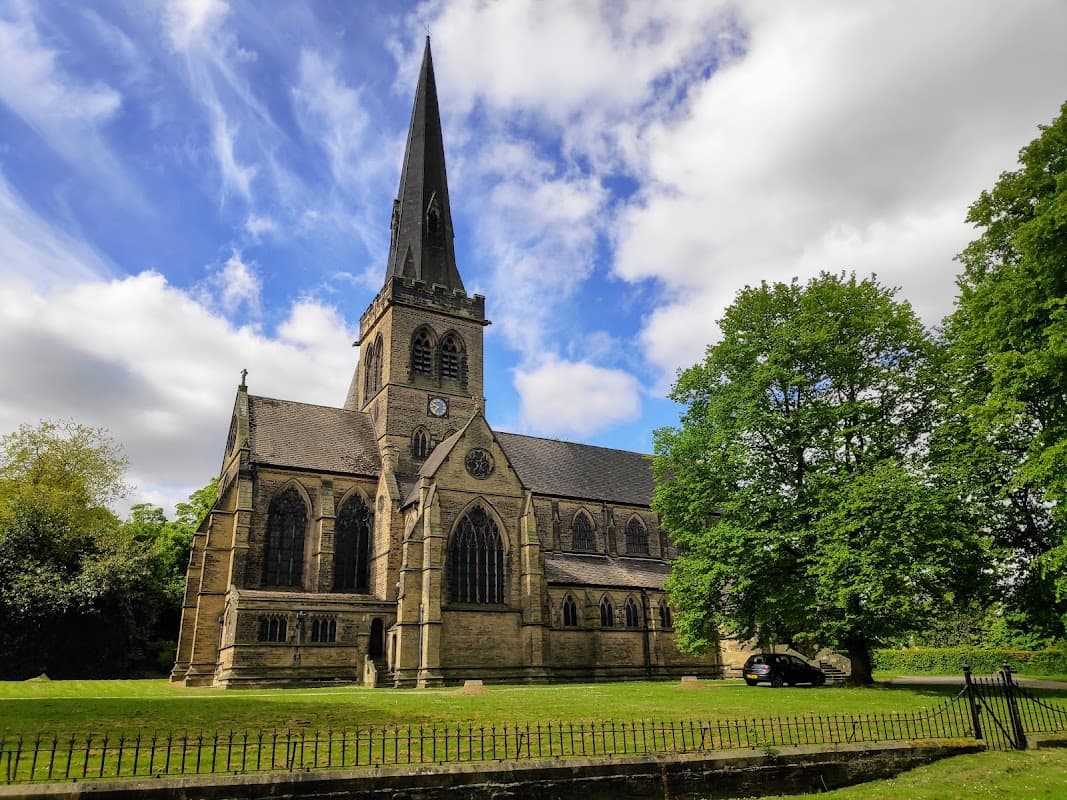 Holy Trinity Church with a tall spire, surrounded by lush greenery and a cloudy blue sky in Wentworth, Yorkshire.