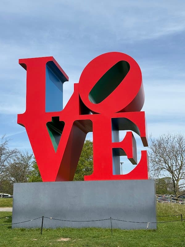 Large red and blue "LOVE" sculpture in a grassy area with trees and a clear blue sky in the background.